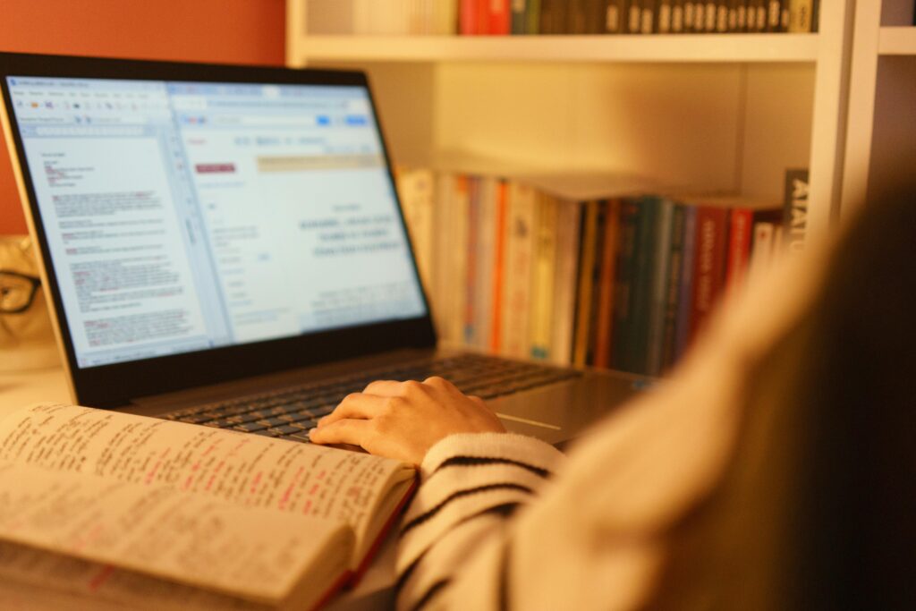 A person studying online with a laptop and open book in a cozy home setting, surrounded by shelves of books.